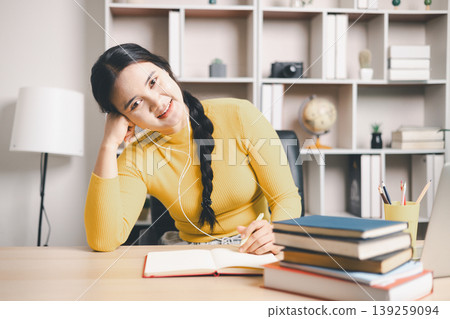 Young Asian woman studying online at home desk, writing notes with earphones and books beside laptop. Smiling student, remote learning, virtual class, education and motivation concept. Young Asian woman studying online at home desk, writing notes with earphones and books beside laptop. Smiling student, remote learning, virtual class, education and motivation concept. 139259094