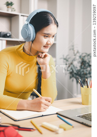 Young Asian woman studying online at home, wearing headphones, writing notes and using laptop at desk. Remote learning, e-learning, focused education, productivity and modern lifestyle concept. Young Asian woman studying online at home, wearing headphones, writing notes and using laptop at desk. Remote learning, e-learning, focused education, productivity and modern lifestyle concept. 139259097