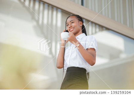 Young African business woman talking on smartphone while holding a cup of coffee, low angle view, concept of mobile communication, remote work and positive modern business lifestyle. Young African business woman talking on smartphone while holding a cup of coffee, low angle view, concept of mobile communication, remote work and positive modern business lifestyle. 139259104