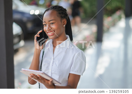 Young African business woman smiling during a phone call while holding tablet outdoors, concept of mobile business, remote communication, digital workflow and flexible work lifestyle. Young African business woman smiling during a phone call while holding tablet outdoors, concept of mobile business, remote communication, digital workflow and flexible work lifestyle. 139259105