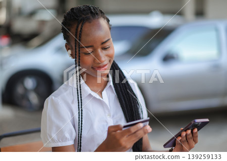 Young African woman holding credit card and smartphone while wearing wireless earbud outdoors, concept of online payment, mobile banking, fintech and secure digital shopping. 139259133
