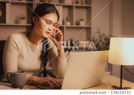 Young Asian woman in glasses working on laptop in cozy home office at night, showing deep focus, remote work, online study, problem solving, concentration and calm evening productivity. 139259135