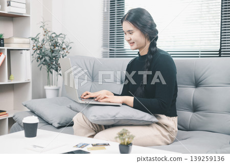 Young Asian woman using laptop on sofa at home for online shopping, browsing ecommerce website with credit cards on table, digital payment, cashless purchase and consumer lifestyle concept 139259136