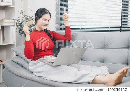 Young Asian woman celebrating online shopping success with laptop and credit card at home, pointing up for sale or cashback offer, ecommerce payment, discount deal and digital purchase concept 139259137