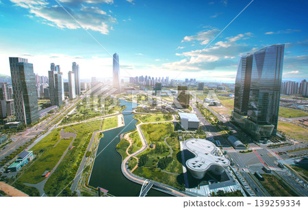 A city skyline shows tall buildings next to a river. The sun shines brightly over the landscape. Green spaces are visible along the water. Roads and paths are nearby. A city skyline shows tall buildings next to a river. The sun shines brightly over the landscape. Green spaces are visible along the water. Roads and paths are nearby. 139259334