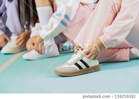 Children are sitting on a blue gym floor tying their shoes before starting a fun activity. They wear casual clothes and look excited to begin their lesson soon. 139259392