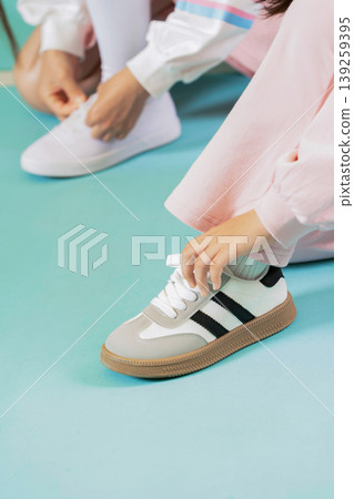 One person is tying the laces of their shoes on a blue floor. Another person sits nearby and is getting ready to put on white sneakers. Both individuals are engaged in their activity. One person is tying the laces of their shoes on a blue floor. Another person sits nearby and is getting ready to put on white sneakers. Both individuals are engaged in their activity. 139259395
