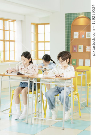 Three students are seated at their desks in a bright classroom. They are focused on their work, writing in notebooks. Sunlight fills the room through the windows. Three students are seated at their desks in a bright classroom. They are focused on their work, writing in notebooks. Sunlight fills the room through the windows. 139259524