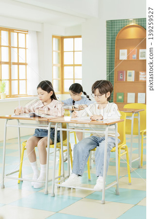 Three children are engaged in writing and studying at their desks in a classroom. Sunlight streams through the windows, illuminating their focused expressions as they work. Three children are engaged in writing and studying at their desks in a classroom. Sunlight streams through the windows, illuminating their focused expressions as they work. 139259527