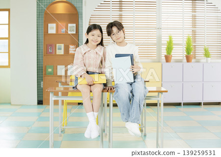 Two children sit on a desk in a classroom during the day. They smile while holding books and show a friendly connection. The room has colorful walls and shelves with books. Two children sit on a desk in a classroom during the day. They smile while holding books and show a friendly connection. The room has colorful walls and shelves with books. 139259531