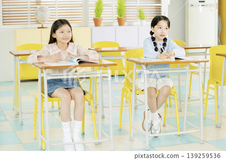 Two young girls are seated at desks in a bright classroom. They appear focused on their work while looking towards the front. The setting is clean and organized with colorful furniture. 139259536