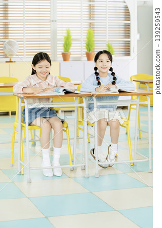 Two girls are sitting at their desks in a classroom. They look happy and are engaged in their activity. The room has bright colors and plants near the windows. Two girls are sitting at their desks in a classroom. They look happy and are engaged in their activity. The room has bright colors and plants near the windows. 139259543