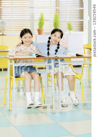 Two girls are sitting at a desk in a classroom. They are smiling and looking happy. One girl waves while the other sits closely and holds a book. The room is filled with light. Two girls are sitting at a desk in a classroom. They are smiling and looking happy. One girl waves while the other sits closely and holds a book. The room is filled with light. 139259549
