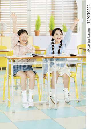 Two girls are seated in a classroom. They are raising their hands, eager to participate in the lesson. The classroom has colorful chairs and sunny decor. 139259551