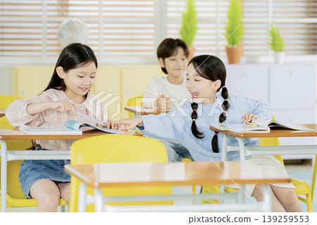 Three children sit at desks in a classroom, focused on learning. Two girls smile and share books while a boy looks on, creating a lively learning environment. Three children sit at desks in a classroom, focused on learning. Two girls smile and share books while a boy looks on, creating a lively learning environment. 139259553