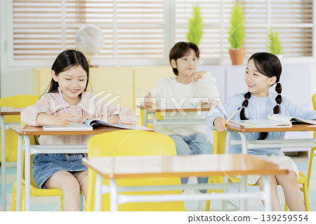 Three children sit at desks in a classroom. They are focused on their books and participate in learning. The room has windows and plants, creating a lively atmosphere. Three children sit at desks in a classroom. They are focused on their books and participate in learning. The room has windows and plants, creating a lively atmosphere. 139259554
