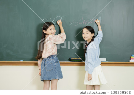 Two girls stand in a classroom writing on a chalkboard. They are preparing to start their school day. Each girl holds a piece of chalk in her hand. Two girls stand in a classroom writing on a chalkboard. They are preparing to start their school day. Each girl holds a piece of chalk in her hand. 139259568