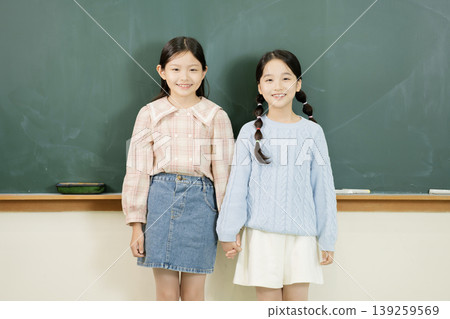 Two girls hold hands and smile in a classroom setting. They wear casual clothes and stand in front of a green chalkboard. The setting shows a typical school environment. 139259569