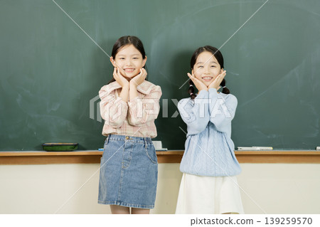 Two girls stand side by side in front of a chalkboard. They smile and hold their hands up to their faces. They wear casual clothes and appear happy during school activities. 139259570