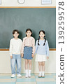 Three children are grouped together in a classroom. Two boys and one girl are holding hands. They are dressed casually and smiling at the camera. 139259578