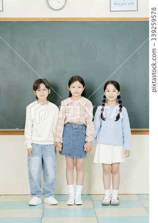 Three children are grouped together in a classroom. Two boys and one girl are holding hands. They are dressed casually and smiling at the camera. Three children are grouped together in a classroom. Two boys and one girl are holding hands. They are dressed casually and smiling at the camera. 139259578