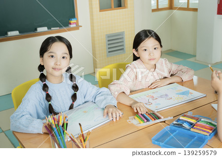 Two girls sit at a classroom table focused on their art projects. They use colored pencils and markers while surrounded by school supplies. The room has bright colors and natural light. Two girls sit at a classroom table focused on their art projects. They use colored pencils and markers while surrounded by school supplies. The room has bright colors and natural light. 139259579
