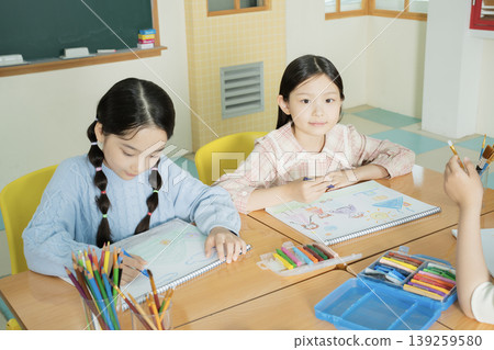 Two children sit at a wooden table in a classroom. They work on drawing with colored pencils and crayons. Bright colors fill their sketchbooks as they focus on their art. Two children sit at a wooden table in a classroom. They work on drawing with colored pencils and crayons. Bright colors fill their sketchbooks as they focus on their art. 139259580