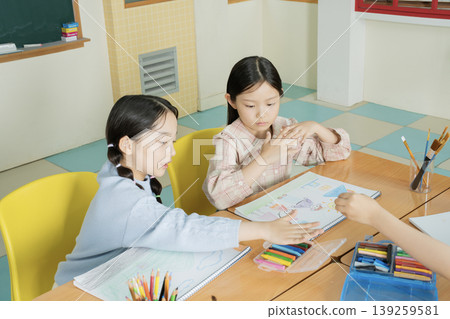 Two children sit at a table in a classroom. One child shows her drawing while the other observes carefully. Art supplies are visible on the table. The room has bright colors. 139259581