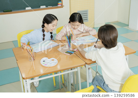 Three children are engaged in drawing and coloring at a table in a classroom. They are using colored pencils and discussing their artwork. The setting shows a bright and friendly learning space. 139259586