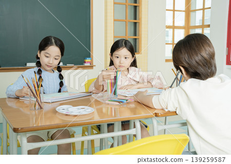 Three children work on their art projects in a classroom. They are seated at a table with colored pencils and sketchbooks. One child holds colored pencils and looks thoughtful. Three children work on their art projects in a classroom. They are seated at a table with colored pencils and sketchbooks. One child holds colored pencils and looks thoughtful. 139259587