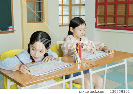 Two children sit at their desks in a classroom. They are focused on coloring pictures with colored pencils. The room has large windows and bright walls. 139259591