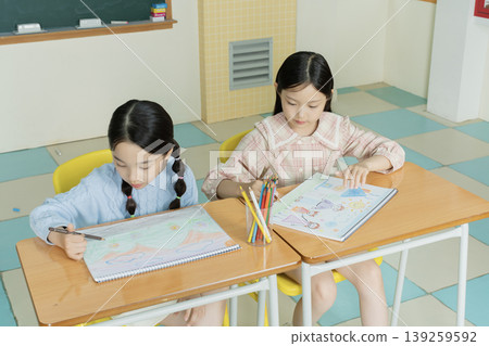Two girls sit at their desks in a classroom and draw on their sketchbooks. They use colored pencils and focus on their artwork. The classroom has colorful tiles and chalkboard. 139259592