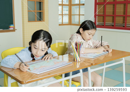 Two young girls focus on their coloring books in a bright classroom. They sit at wooden desks and use colored pencils to create their art. The room has large windows and colorful tiles. 139259593