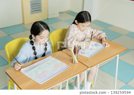Two children sit at desks in a classroom, focused on their drawings. They use colored pencils to create colorful designs in their sketchbooks during an art session. 139259594