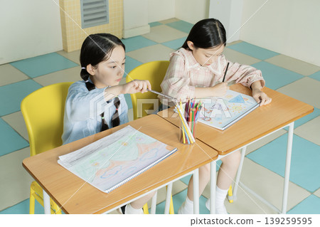 Two girls sit at separate desks engaged in a drawing and coloring activity. They use colored pencils to create their artwork, focused on their projects. Bright colors fill the room. 139259595