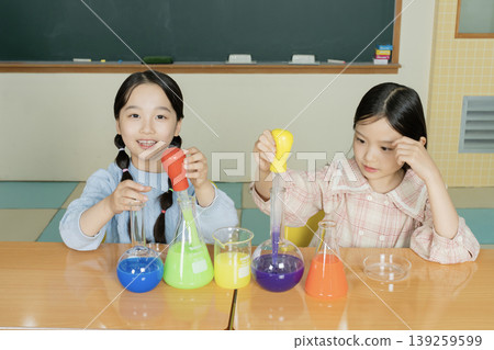 Two girls engage in a science activity by mixing colorful liquids in beakers. The classroom is bright with a chalkboard in the background. They smile and focus on their experiment. 139259599