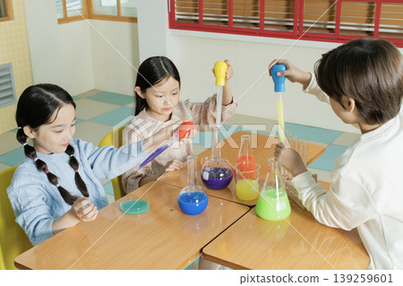 Three kids are working together at a table in a classroom. They are mixing different colored liquids into glass flasks. Each child uses a dropper or a container to add to the flasks. Three kids are working together at a table in a classroom. They are mixing different colored liquids into glass flasks. Each child uses a dropper or a container to add to the flasks. 139259601
