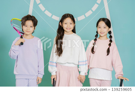Three young children stand together on a tennis court. They hold rackets and smile. The background is a bright blue with a tennis net. It looks like they enjoy the game and have fun. Three young children stand together on a tennis court. They hold rackets and smile. The background is a bright blue with a tennis net. It looks like they enjoy the game and have fun. 139259605