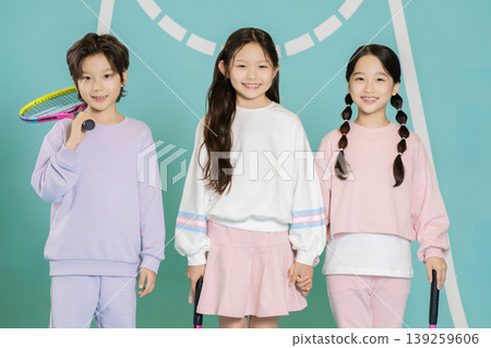 Three children stand together at a tennis court. They hold tennis rackets and wear matching outfits. They are smiling and look happy. The setting is bright and colorful, showing a playful mood. Three children stand together at a tennis court. They hold tennis rackets and wear matching outfits. They are smiling and look happy. The setting is bright and colorful, showing a playful mood. 139259606
