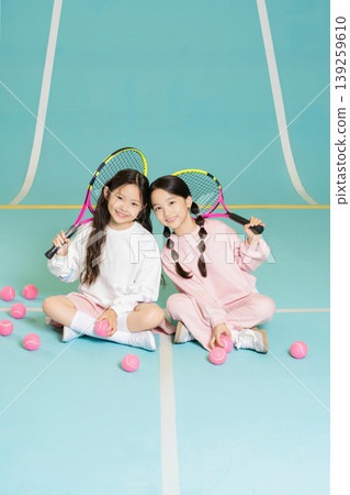 Two young girls sit on a blue indoor tennis court. They hold tennis rackets and smile. Pink tennis balls are scattered around them. They appear happy and ready to play tennis. 139259610