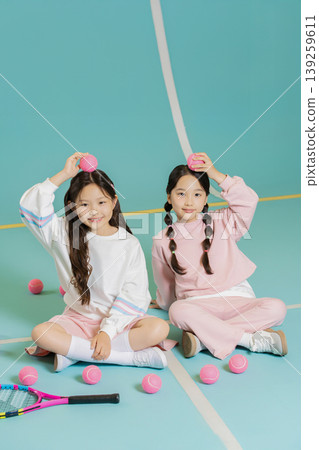 Two girls sit on a court and hold pink balls above their heads. They smile while surrounded by more pink balls and a tennis racket, enjoying their time together in a playful moment. 139259611