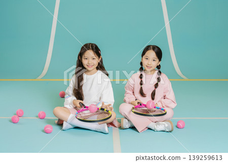 Two girls are seated on the gym floor, each holding a tennis racket. They are surrounded by pink tennis balls. The location has a light blue background, creating a bright and playful setting. Two girls are seated on the gym floor, each holding a tennis racket. They are surrounded by pink tennis balls. The location has a light blue background, creating a bright and playful setting. 139259613