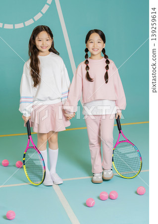 Two girls stand together on a tennis court, each holding a tennis racket. They smile at the camera while surrounded by tennis balls. They wear comfortable sports clothing in bright colors. Two girls stand together on a tennis court, each holding a tennis racket. They smile at the camera while surrounded by tennis balls. They wear comfortable sports clothing in bright colors. 139259614