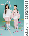 Two young girls hold tennis rackets while standing on a court. They are smiling at the camera, with pink tennis balls around them. They wear casual sports clothes and look happy. 139259615