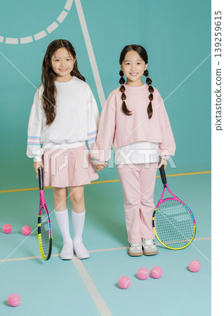 Two young girls hold tennis rackets while standing on a court. They are smiling at the camera, with pink tennis balls around them. They wear casual sports clothes and look happy. Two young girls hold tennis rackets while standing on a court. They are smiling at the camera, with pink tennis balls around them. They wear casual sports clothes and look happy. 139259615