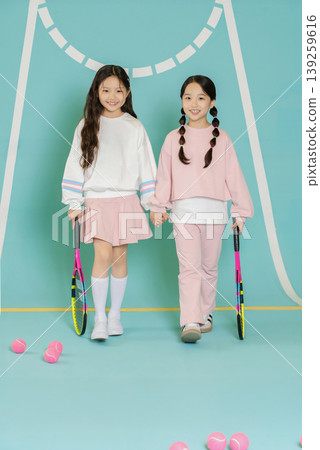 Two young girls walk together on a colored tennis court. They hold tennis rackets and smile while surrounded by pink tennis balls and colored backgrounds. Two young girls walk together on a colored tennis court. They hold tennis rackets and smile while surrounded by pink tennis balls and colored backgrounds. 139259616