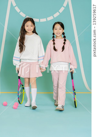 Two girls are walking side by side in a sports area. Both hold tennis rackets and wear friendly smiles. They wear comfortable sports outfits and stand against a colorful background. Two girls are walking side by side in a sports area. Both hold tennis rackets and wear friendly smiles. They wear comfortable sports outfits and stand against a colorful background. 139259617
