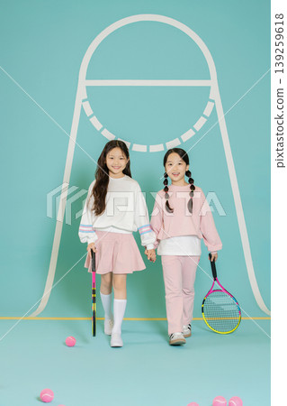 Two girls hold hands while walking on a colorful tennis court. They wear matching sports outfits and smile. Tennis rackets and balls are scattered on the ground. Two girls hold hands while walking on a colorful tennis court. They wear matching sports outfits and smile. Tennis rackets and balls are scattered on the ground. 139259618
