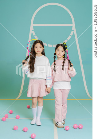 Two young girls stand side by side on a tennis court. They hold tennis rackets and smile at the camera. Pink balls scatter around them on the floor. The court background is simple. Two young girls stand side by side on a tennis court. They hold tennis rackets and smile at the camera. Pink balls scatter around them on the floor. The court background is simple. 139259619