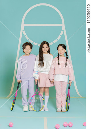 Three children stand together on a tennis court holding rackets. They are dressed in comfortable clothes while surrounded by tennis balls and pink sports items. The background features playful colors. Three children stand together on a tennis court holding rackets. They are dressed in comfortable clothes while surrounded by tennis balls and pink sports items. The background features playful colors. 139259620